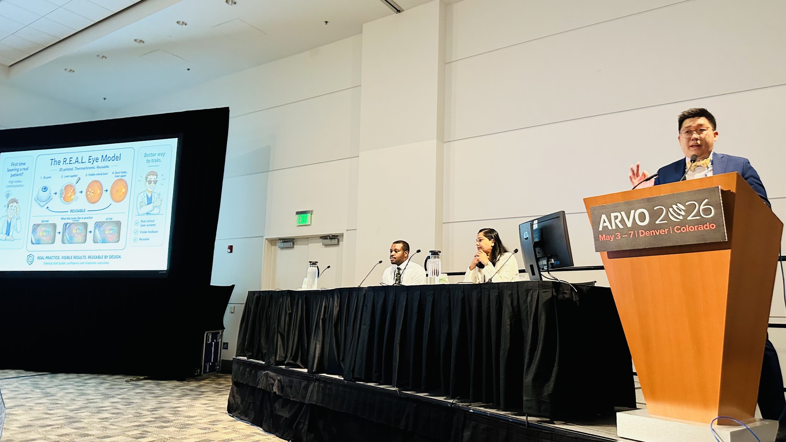 A man in a blue blazer is giving a presentation at a conference called ARVO 2026, held in Denver, Colorado. There is a large presentation slide on the screen titled "The R.E.A.L. Eye Model" with diagrams and text.