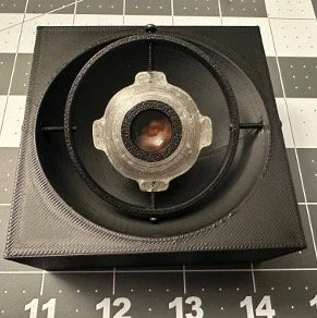 Close-up of a camera lens mounted on a black case or box, with a grid-patterned background.
