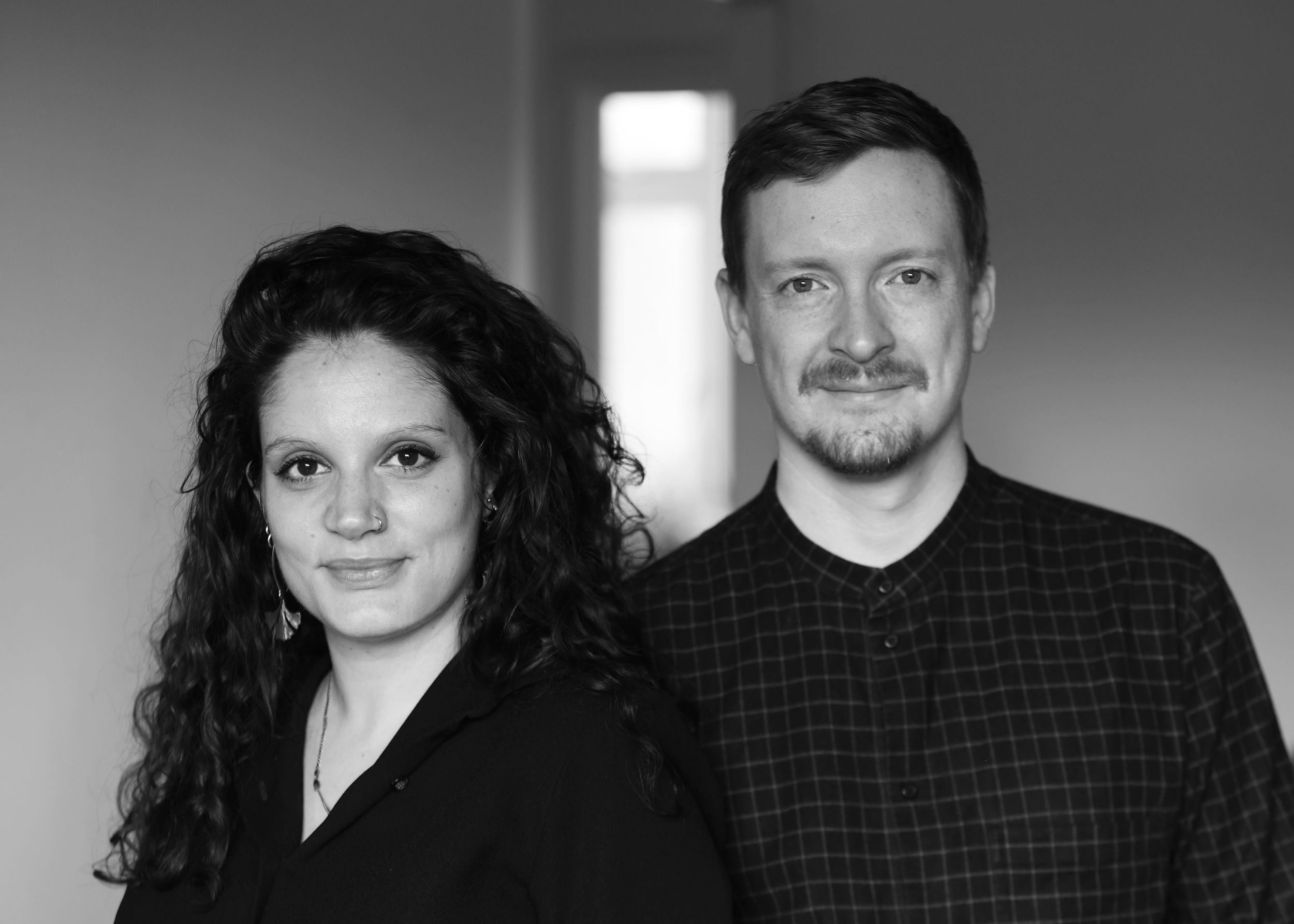 A black and white photograph of a man and woman standing side by side indoors, with a window in the background. The woman has curly hair, a nose piercing, and is wearing earrings and a dark top. The man has short hair, a light beard, and is wearing a checkered shirt.