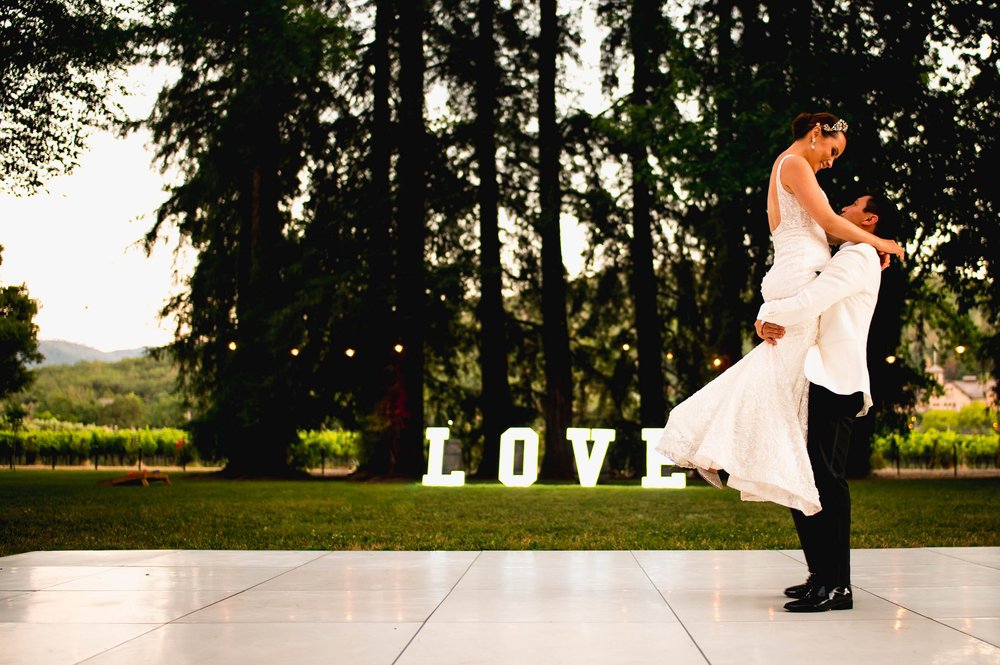 groom holding his bride up in the air as they share their first dance in front of their neon love sign