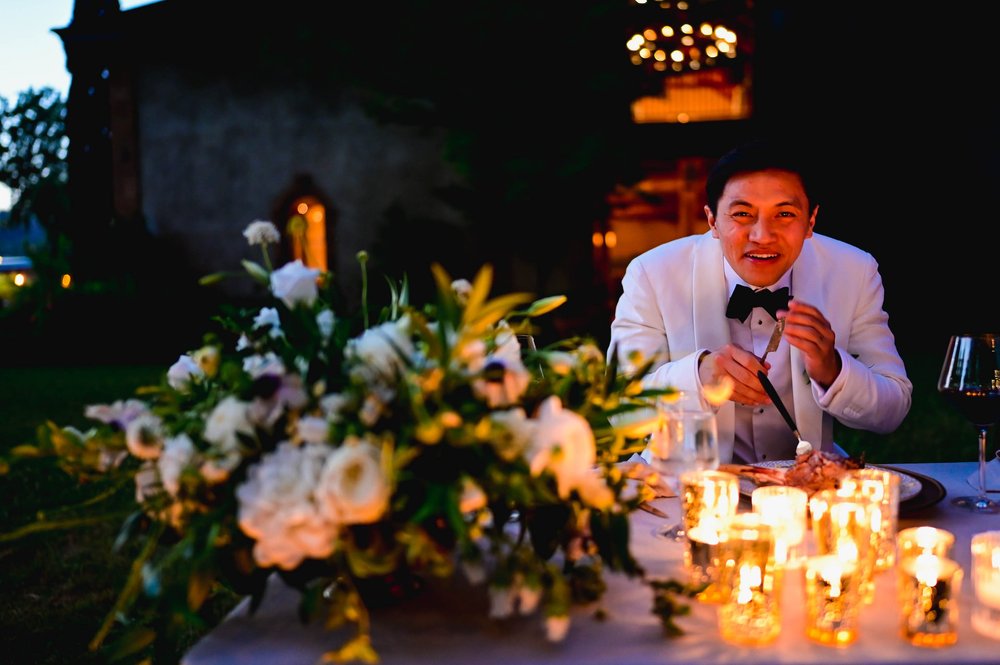 the groom sneaks a bite from his meal and laughs as he is caught by the photographer