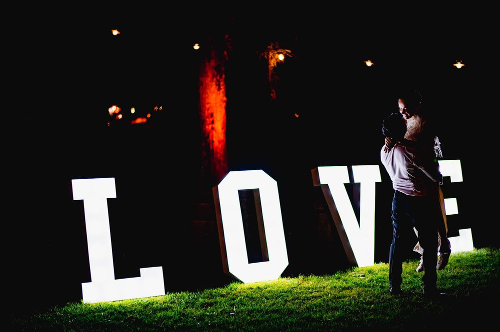 the groom holds up the bride in the grass by their neon love sign in the dark