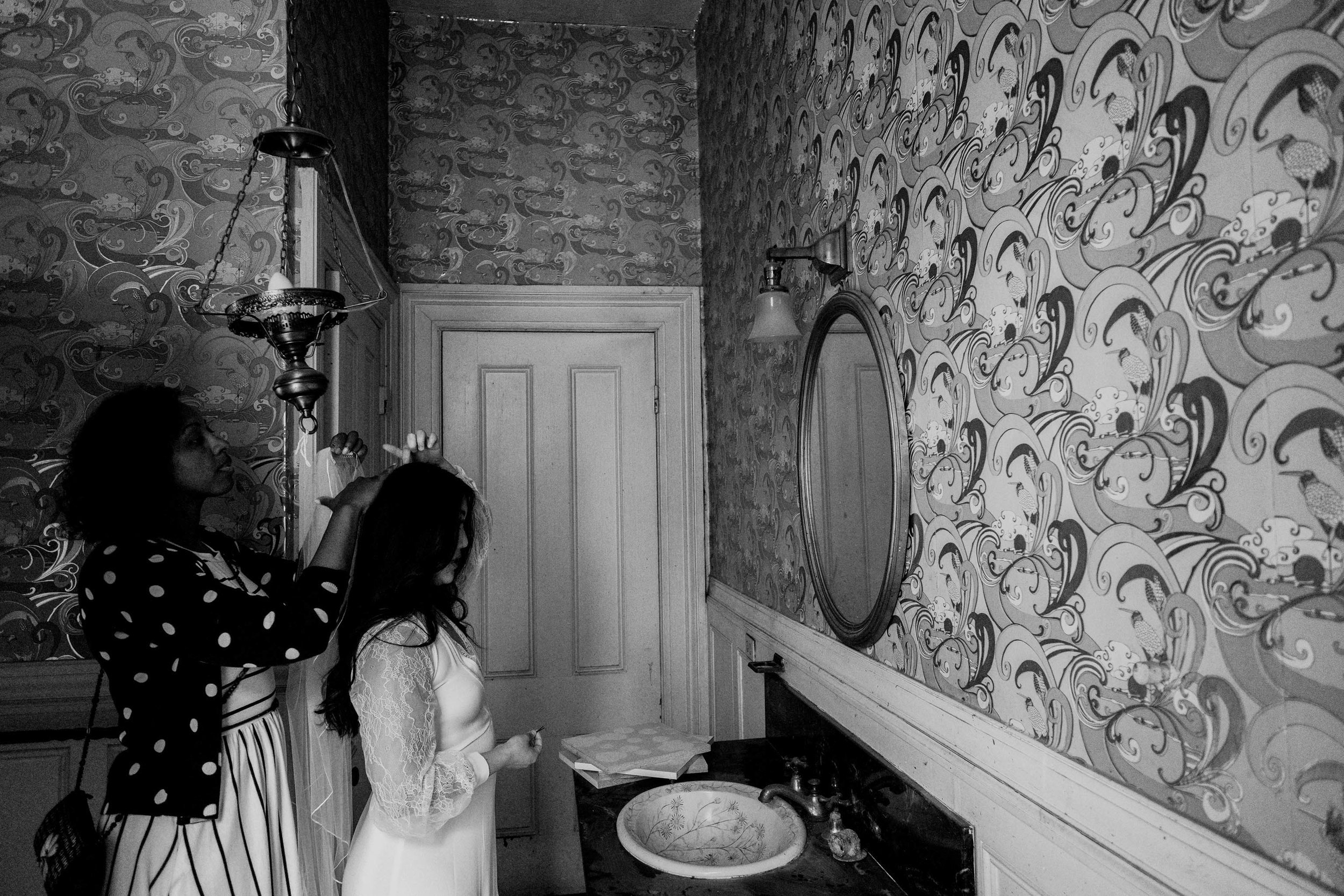 Bride and best friend getting bride ready with her veil in the bathroom mirror