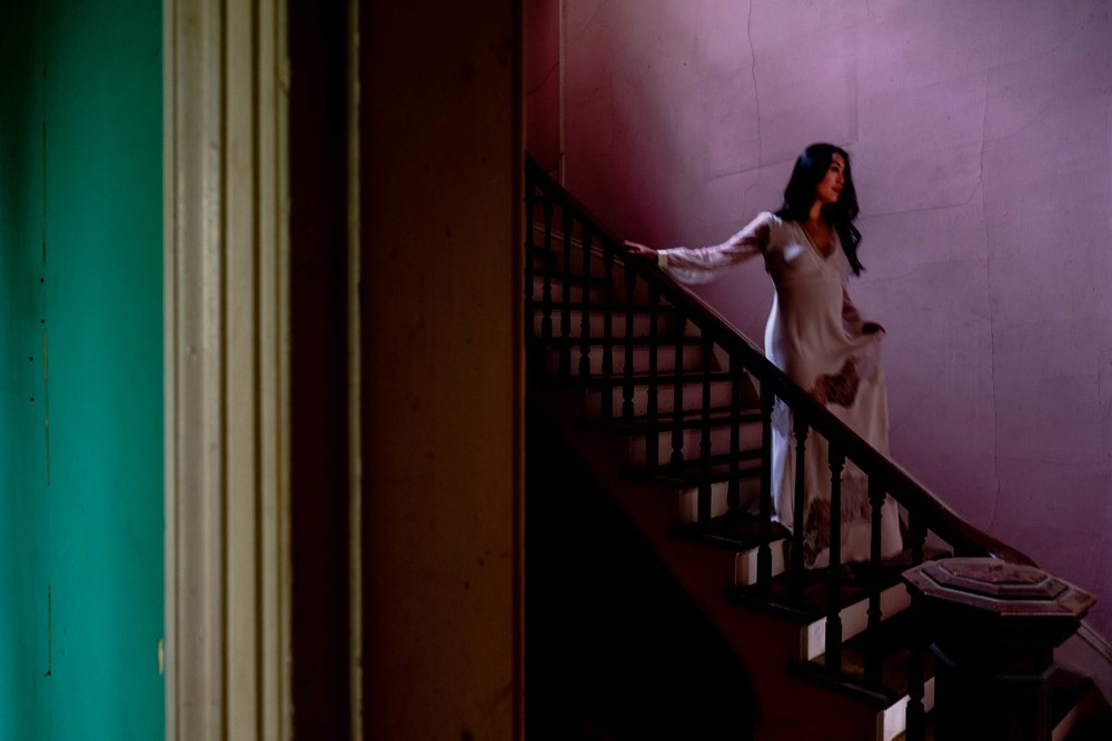 Bride standing on the stairs with bright pink wall behind her and bright green wall on the wall through the doorway