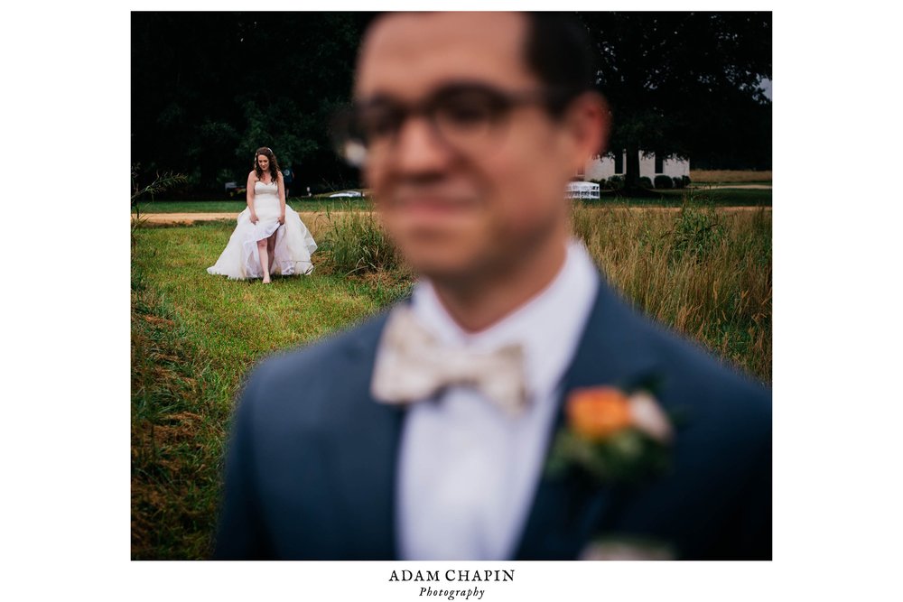 groom blurred out in the foreground as his bride is walking up from behind though the field for their first look