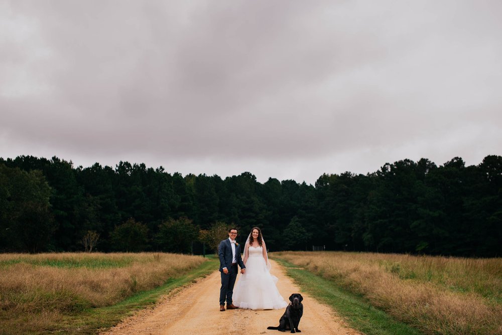 bride and groom standing in the drive of the Meadows Raleigh with their black labrador retriever sitting in front of them