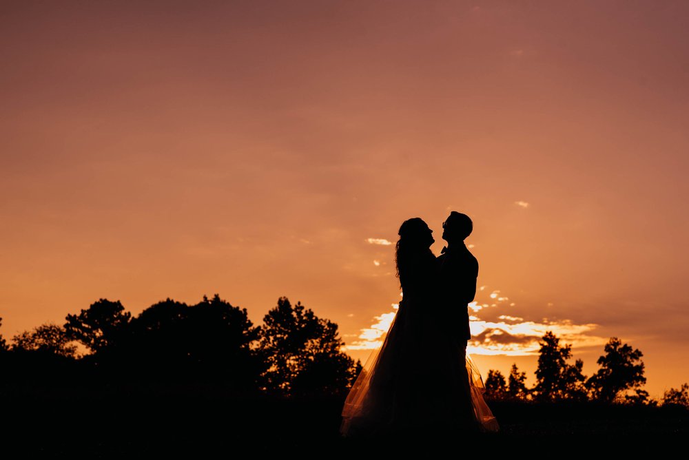 bride and groom standing in the pink and orange sunset as they are silhouetted by the sun