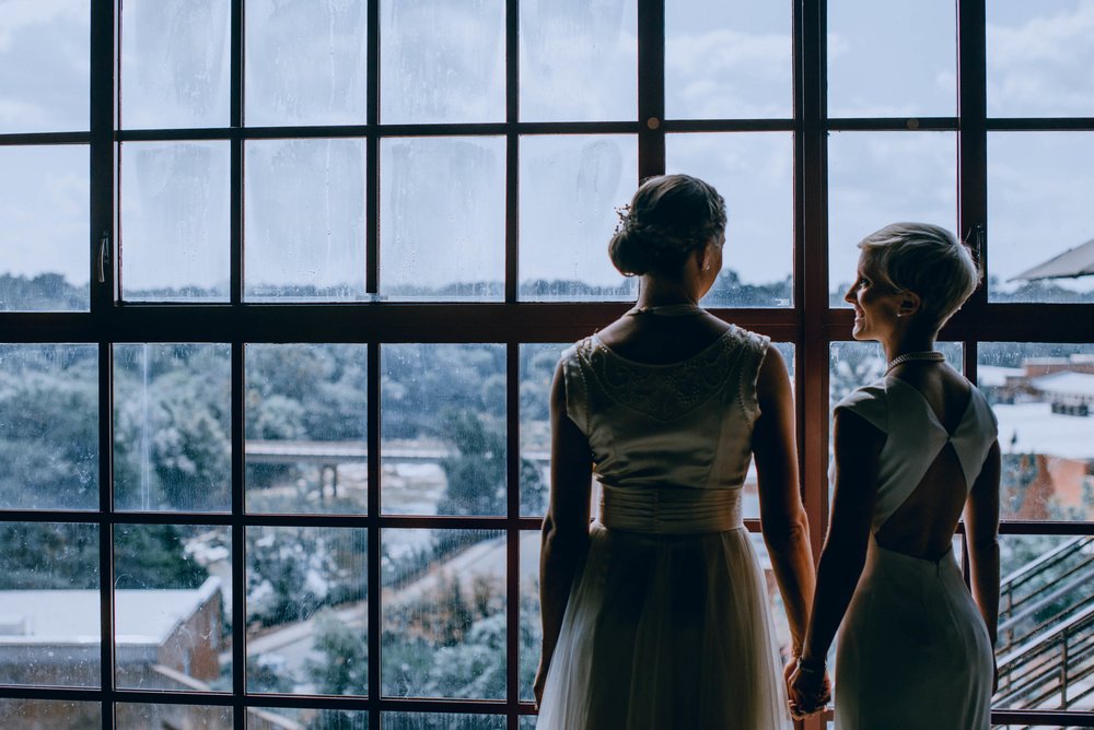 brides looking out the window from the haw river ballroom in saxapahaw north carolina