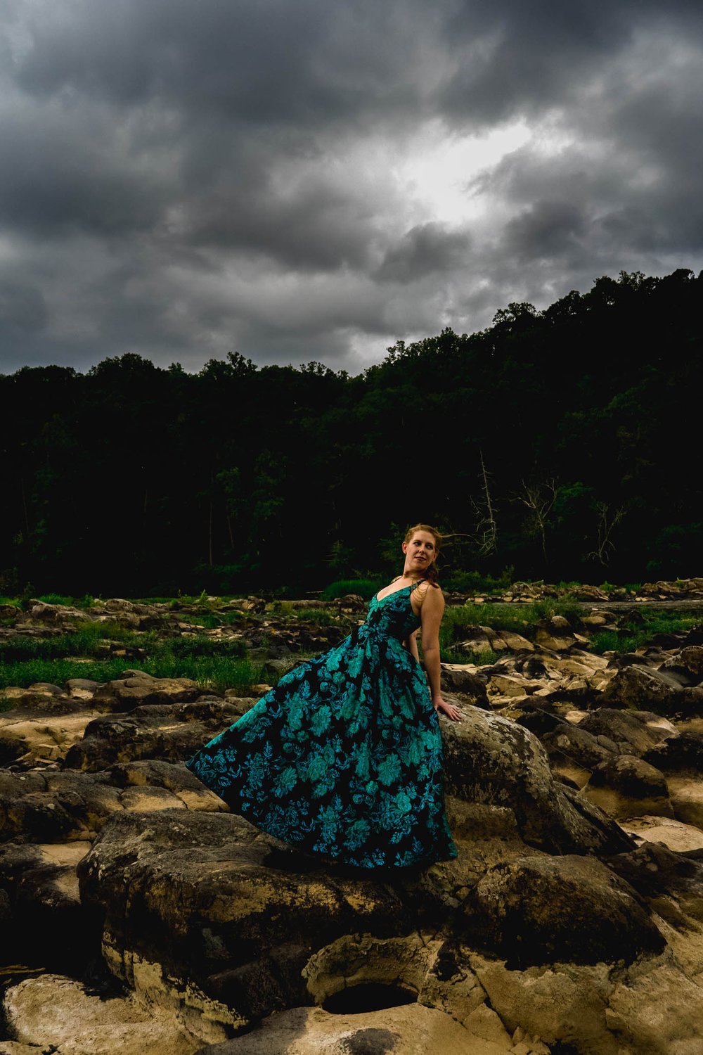 woman poses and tosses her dress next to a rock on a river