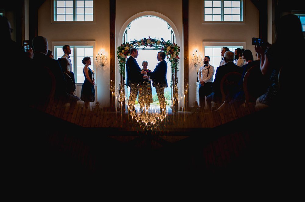 two grooms hold hands during their wedding ceremony as the chandelier is reflected around them