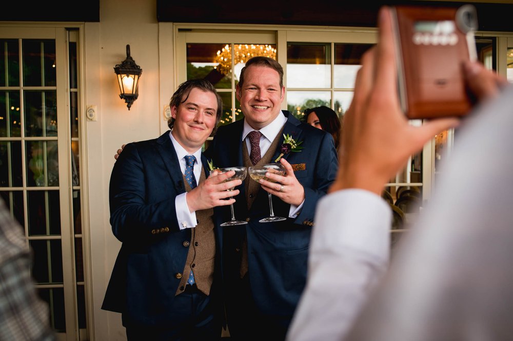 two grooms pose for a polaroid with drinks in hand