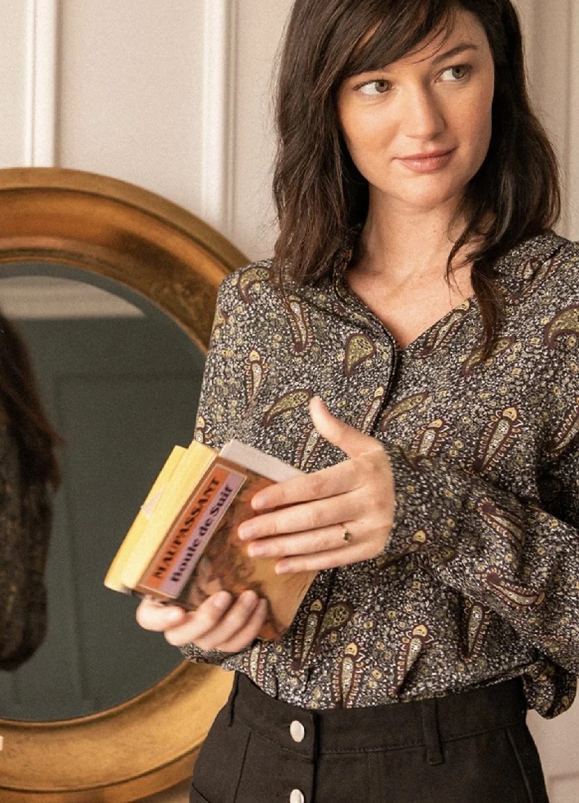 Une femme tenant un livre ou un carnet au format de poche, portant une chemise à motifs et des pantalons noirs, dans une pièce avec un miroir rond en bois en arrière-plan.