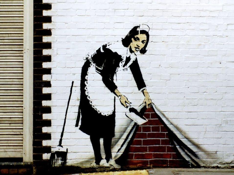 A stencil mural of a uniformed maid lifting the edge of a white curtain or wall and sweeping dust beneath it.