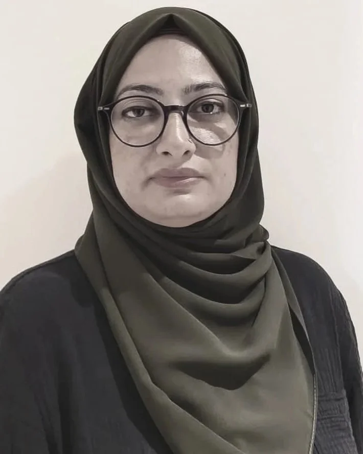 A woman wearing glasses and a dark headscarf standing against a plain white wall.