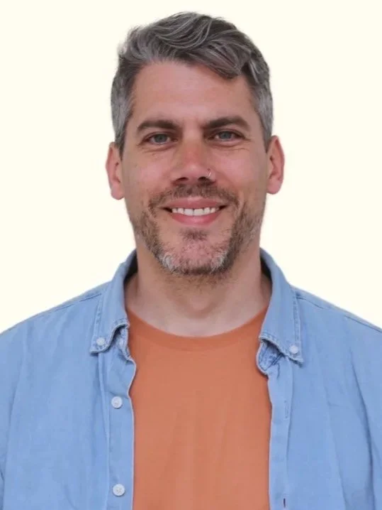 A man with short gray hair, a beard and mustache, wearing a light blue denim shirt over an orange T-shirt, smiling at the camera.