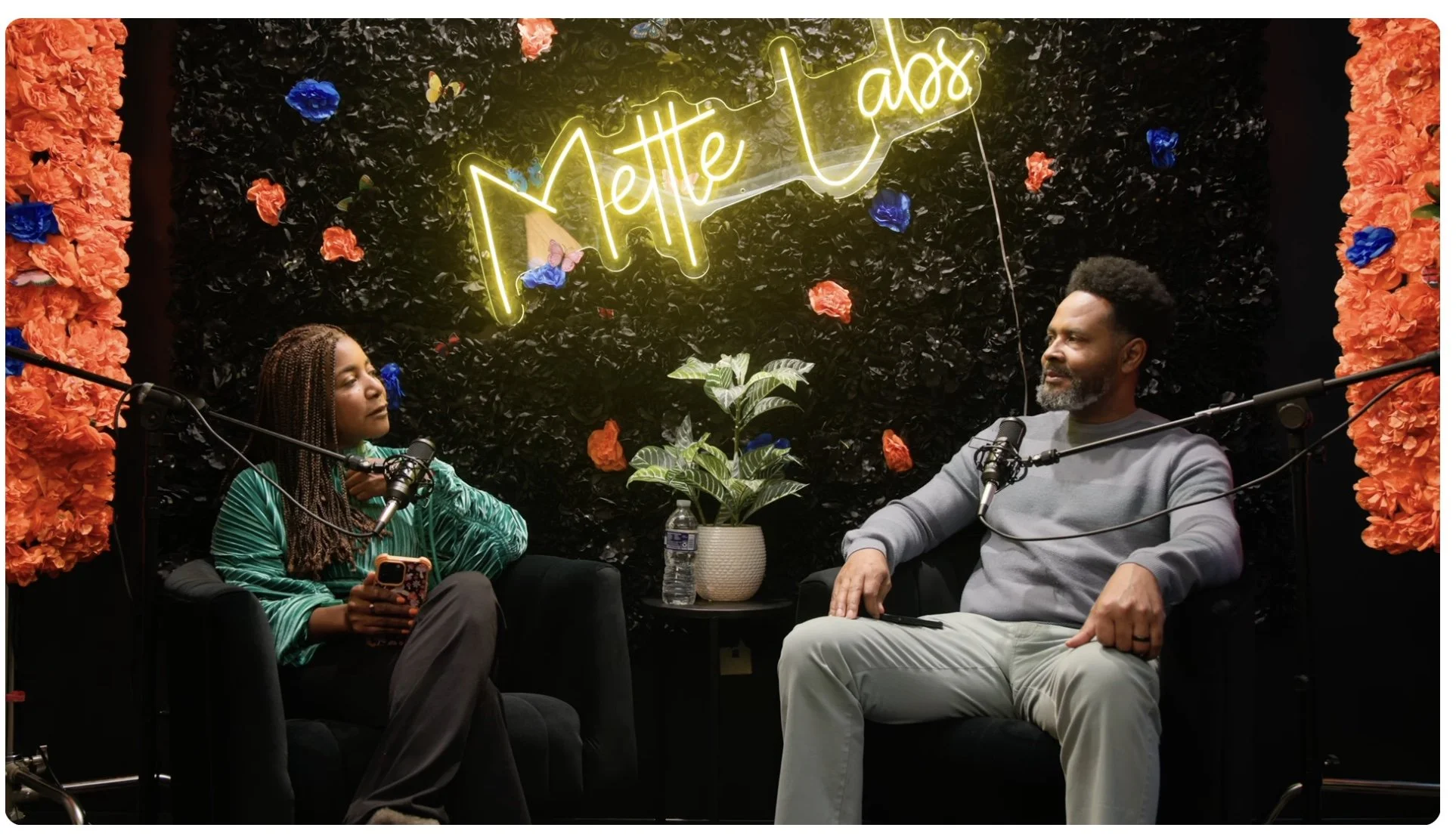 Two people sitting in black armchairs with microphones, having a conversation, in front of a black wall decorated with flowers and a neon sign that reads 'Mettle TV abs'. A potted plant on a small table between them, with a water bottle and a phone. Orange and blue flowers on either side of the scene.