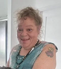 A smiling middle-aged woman with curly blonde hair wearing a sleeveless top and necklaces, standing indoors.