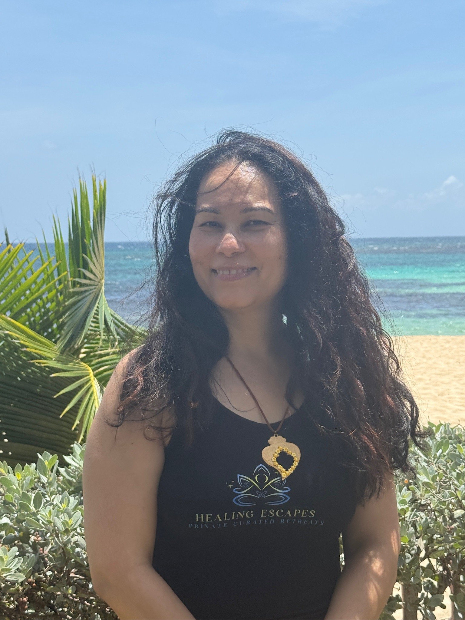 A woman with long, curly dark hair standing on a beach near green plants, with the ocean and blue sky in the background. She is wearing a black tank top with the logo and text "Healing Escapes Private Curated Retreats" and a gold necklace with a heart-shaped pendant.