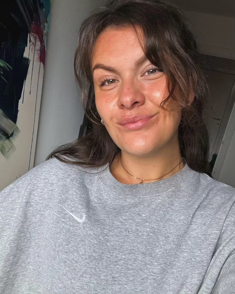 A young woman with light skin, brown wavy hair, and subtle makeup, smiling slightly, wearing a gray Nike sweatshirt, gold necklaces, and small hoop earrings, in a room with a painting or poster and a door in the background.