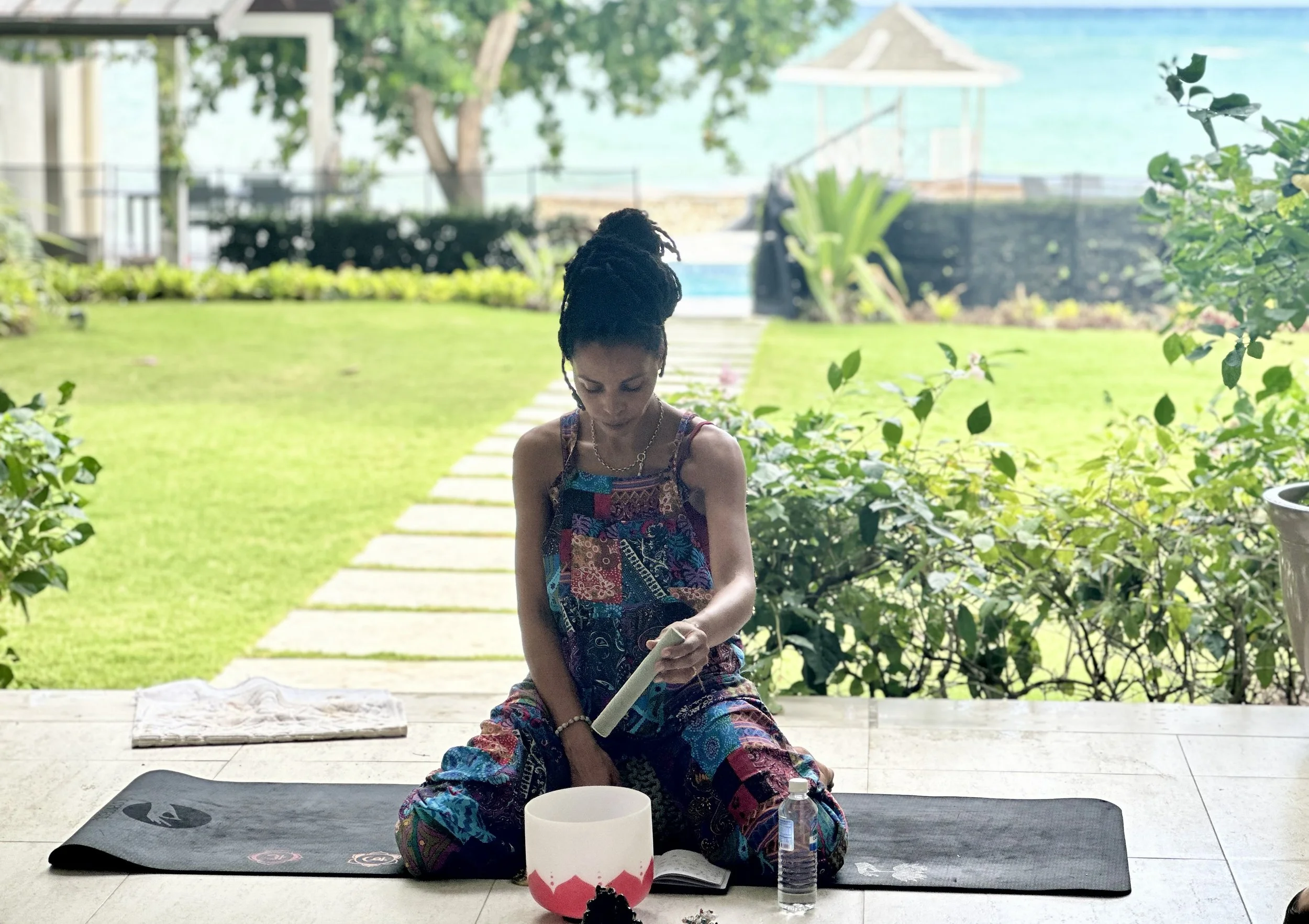 A woman with dark skin and dreadlocks sitting on a yoga mat outdoors, engaging in a meditation practice with a crystal singing bowl, a water bottle, and an open book nearby, with a lush green garden and a view of the ocean in the background.