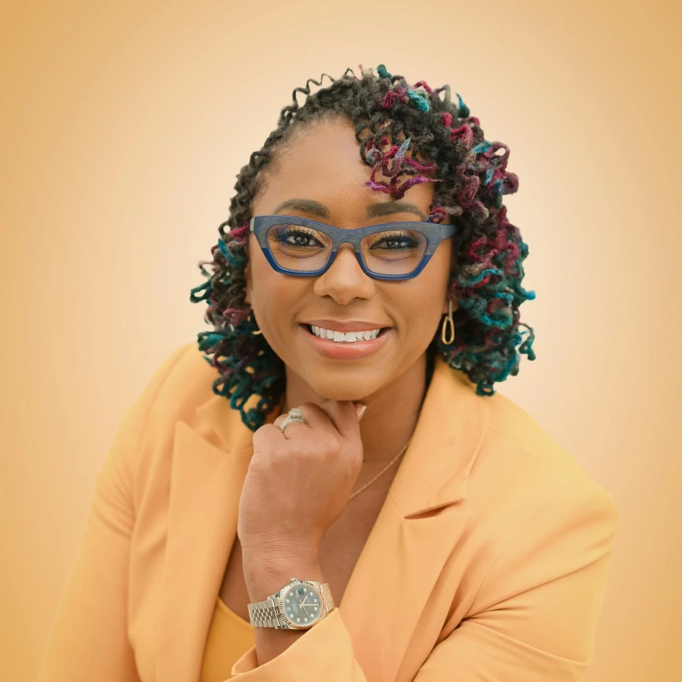 A woman with colorful curly hair, blue glasses, gold earrings, and a watch, smiling and poses with her hand near her chin, wearing a yellow blazer.
