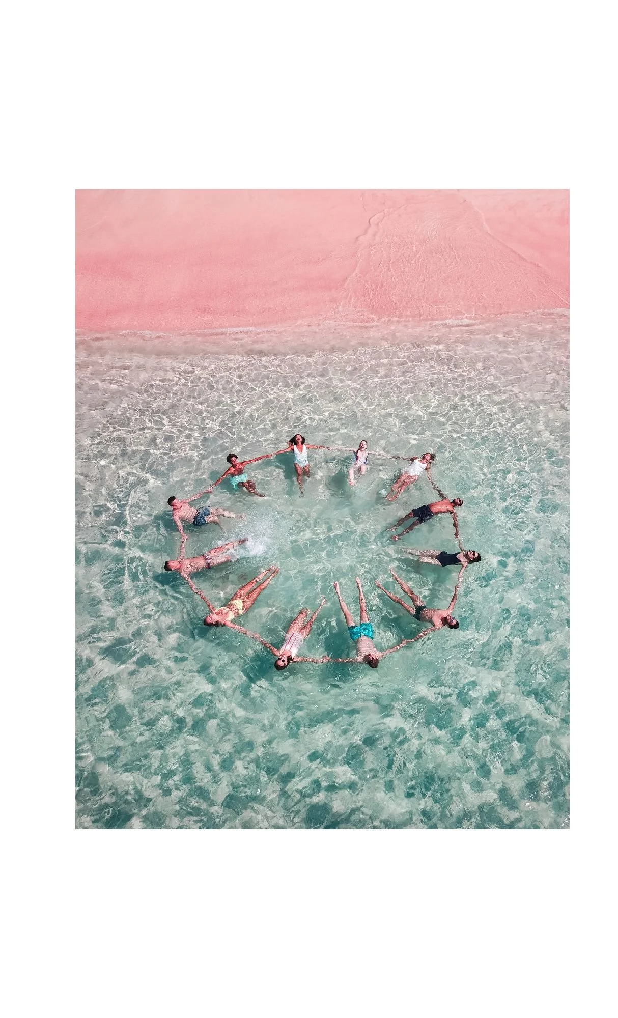 People forming a circle while floating and holding hands in shallow ocean water near pink sandy beach.