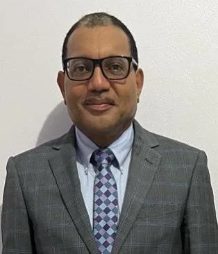 A man with glasses in a gray plaid suit, white shirt, and patterned tie standing against a plain white wall.