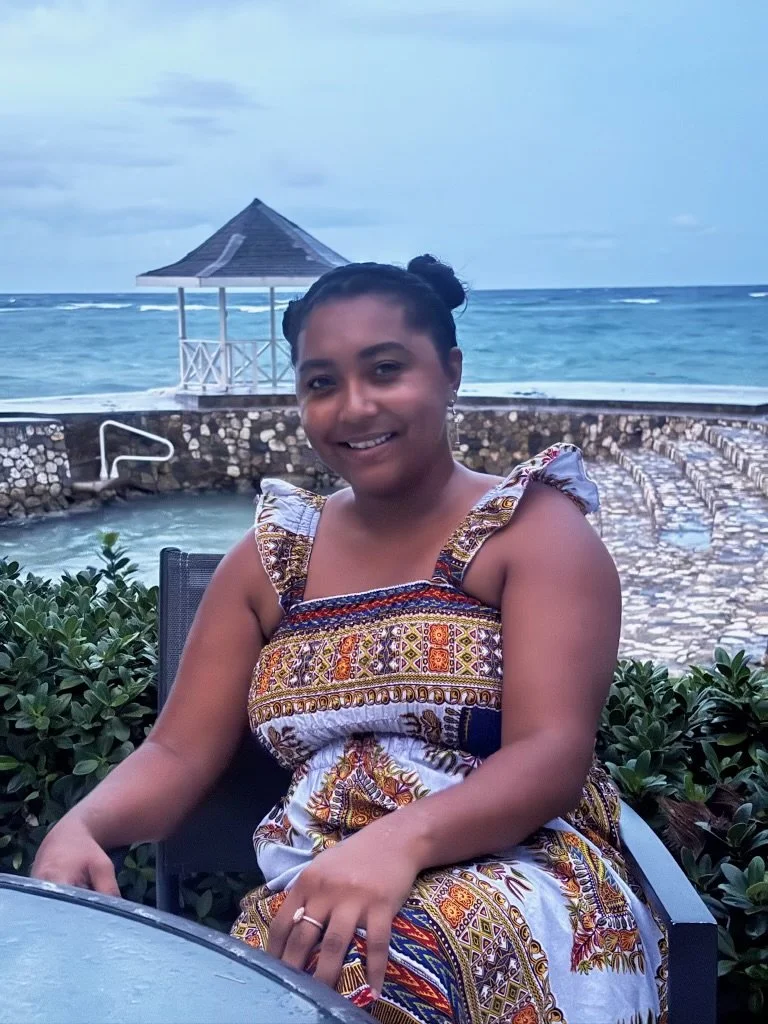 A woman with dark hair styled in two buns, smiling and wearing a colorful, patterned sundress, seated outdoors near a beach with the ocean, a stone pool, and a gazebo in the background.