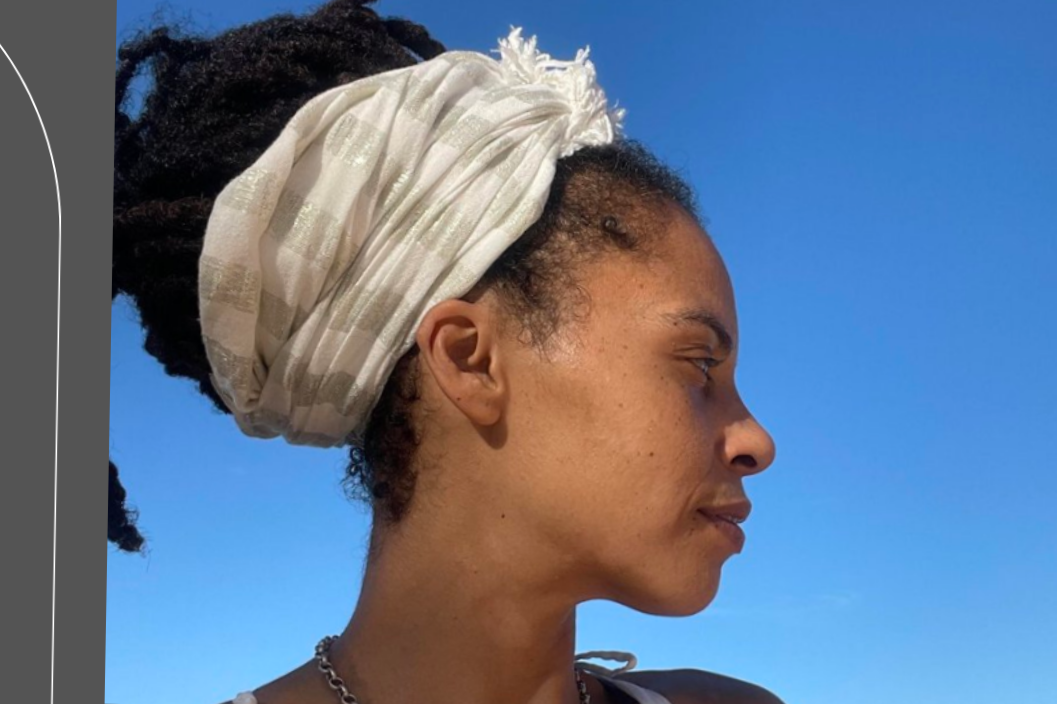 A woman with dark skin and dreadlocks sitting on a yoga mat outdoors, engaging in a meditation practice with a crystal singing bowl, a water bottle, and an open book nearby, with a lush green garden and a view of the ocean in the background.