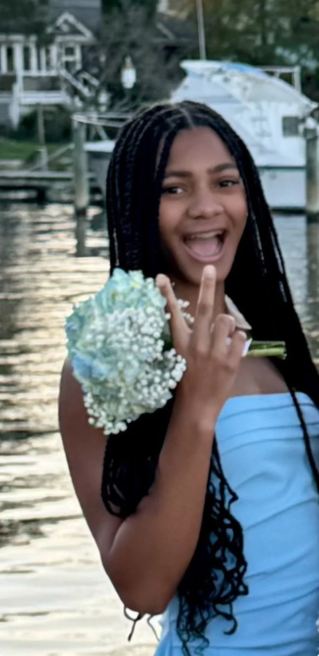 A young woman with long braided hair smiling and showing a peace sign with her fingers, holding a bouquet of white flowers, standing outdoors near water with boats and a house in the background.