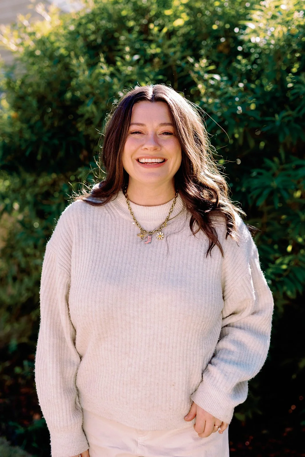 A woman with long brown hair smiling outdoors in front of green foliage, wearing a cream-colored sweater and layered necklaces.