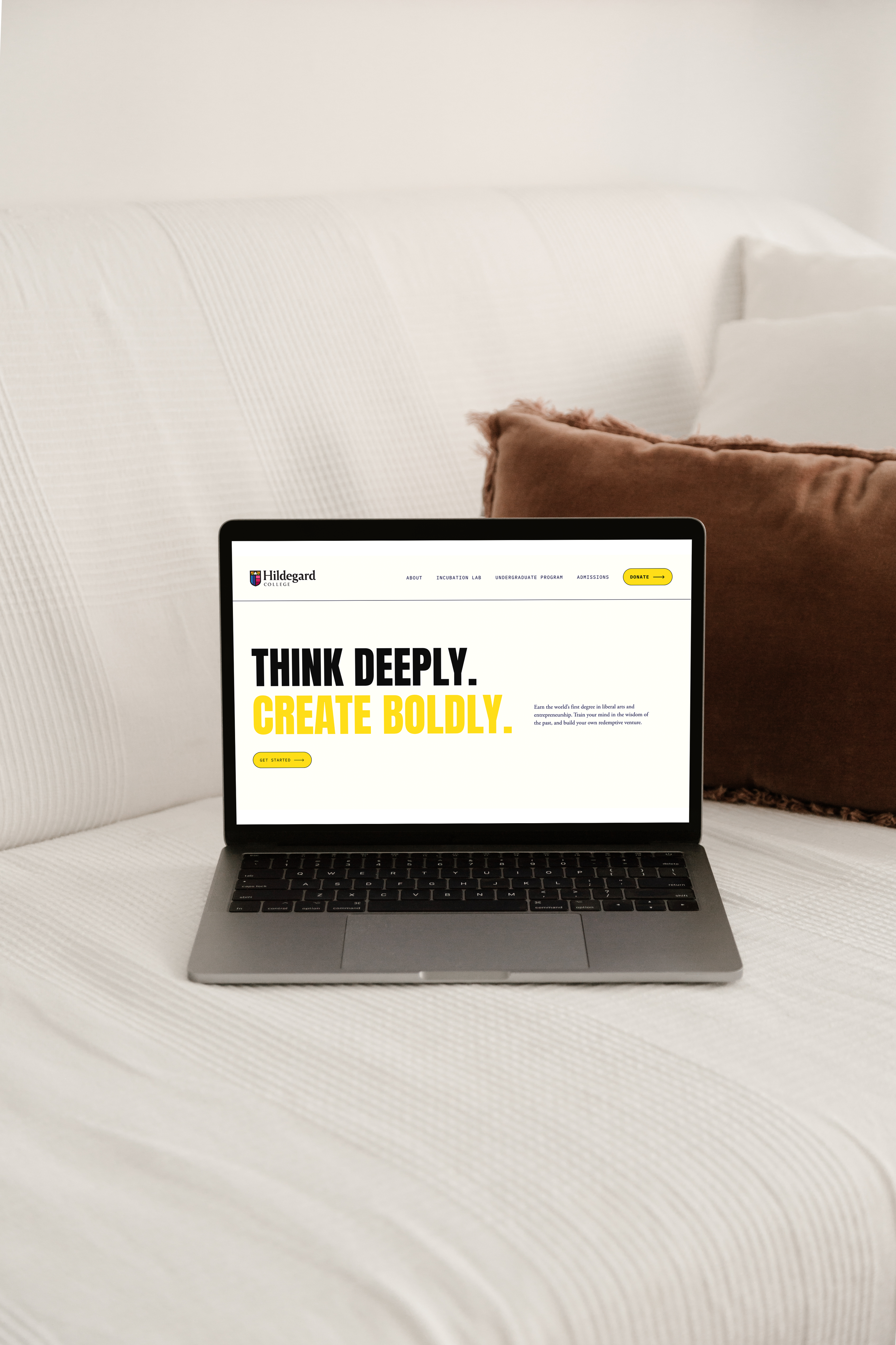 A laptop sitting on a white upholstered bed with pillows, displaying a website with the text 'THINK DEEPLY. CREATE BOLDLY.' in large black and yellow letters.