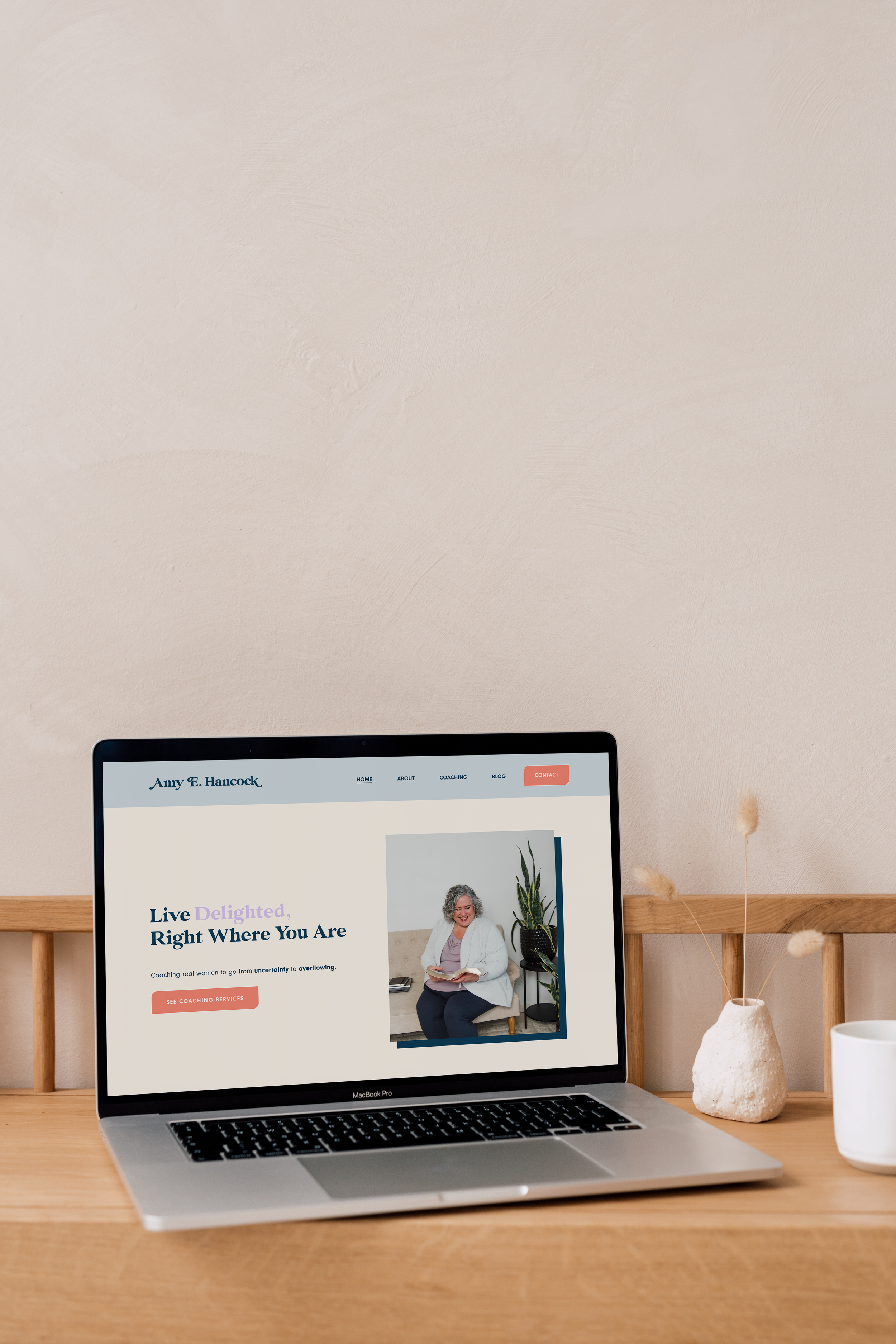 A laptop on a wooden desk displaying a coaching website with the headline "Live Delighted, Right Where You Are" and an image of a smiling woman reading a book. Next to the laptop is a concrete vase with dried flowers and a white cup.