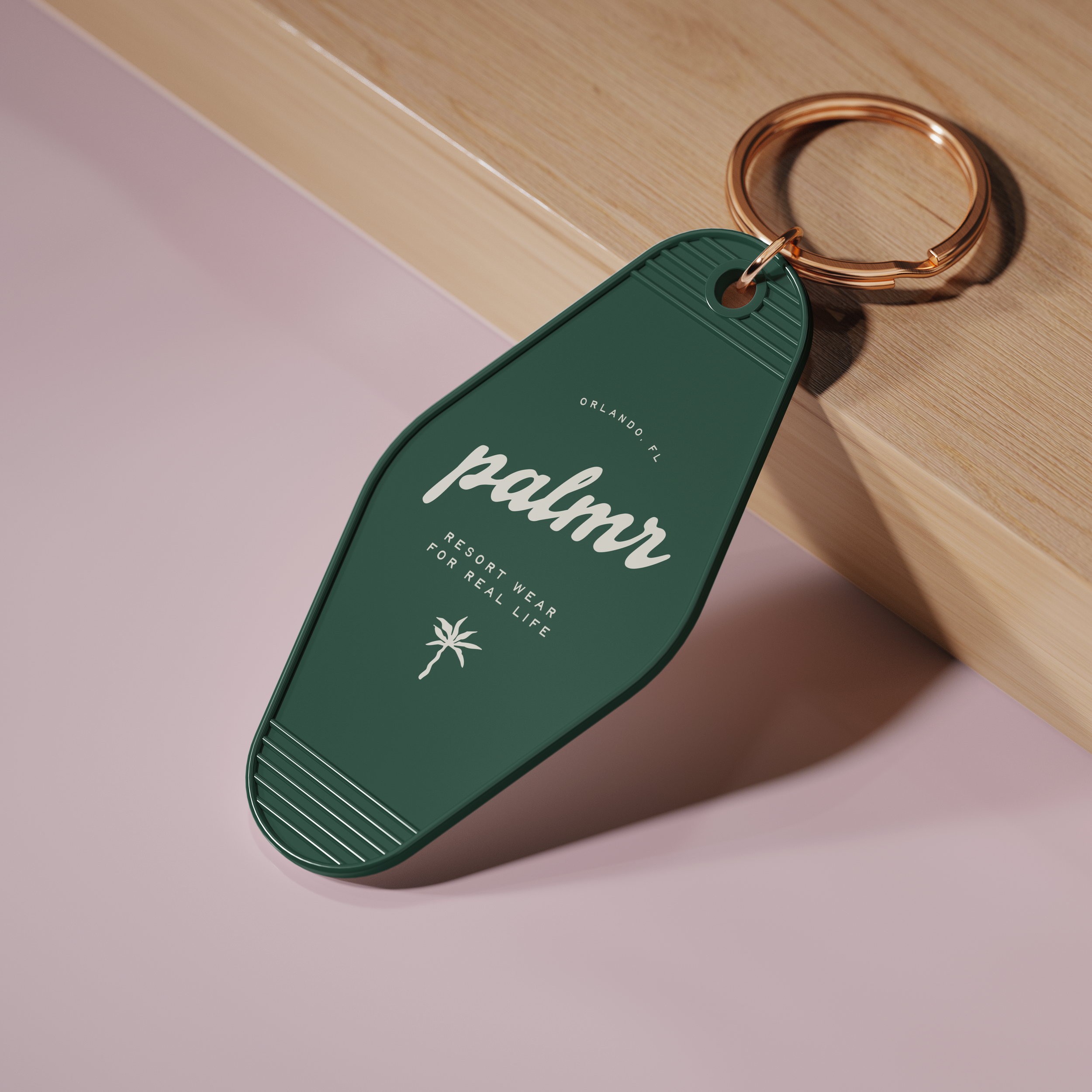 A close-up of a green keychain with white text reading 'Palm' attached to a copper-colored key ring, resting on a wooden surface with a pinkish background.
