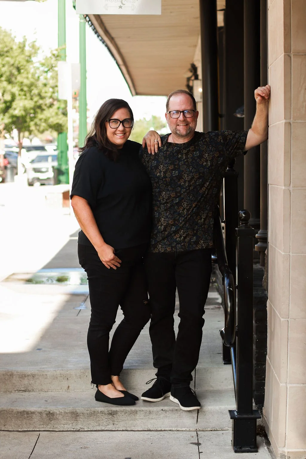 A woman and a man standing outside on a sidewalk, smiling at the camera. The woman is wearing glasses, a black top, black pants, and black flats. The man is wearing glasses, a black patterned shirt, and black pants, with his left arm raised and resting against the wall.
