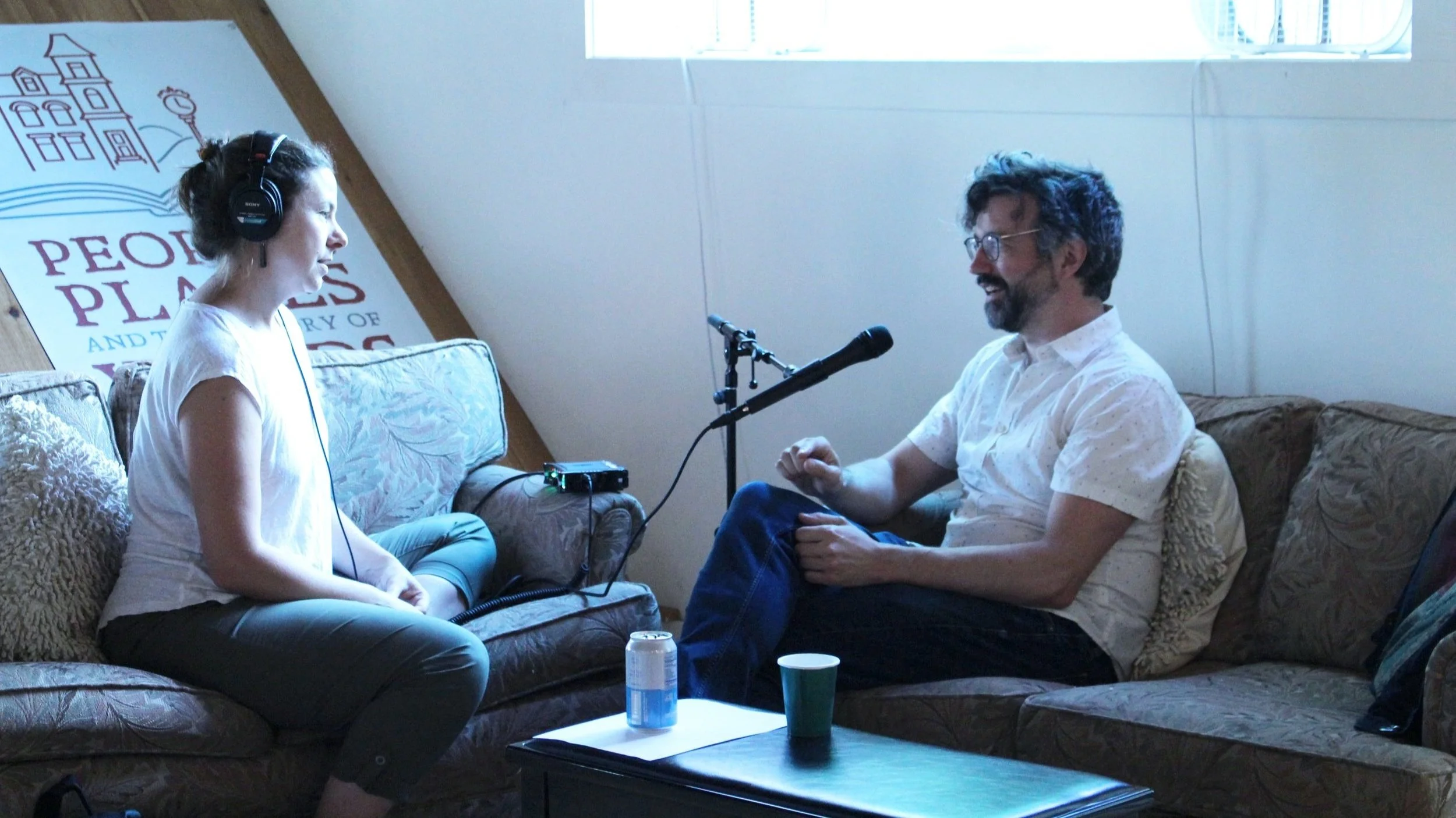 A woman wearing headphones interviews a man in glasses using a microphone and recorder.