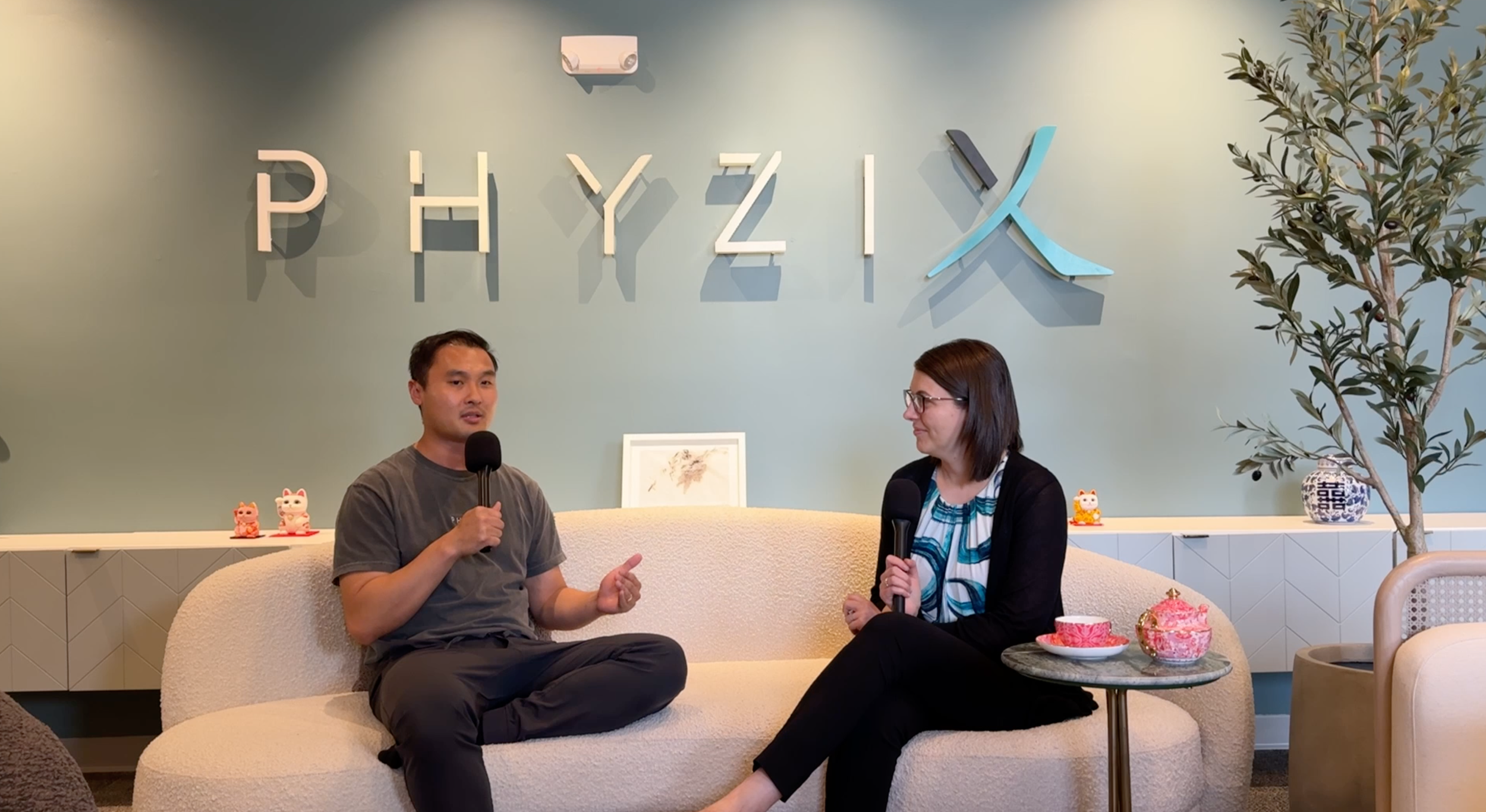 Two people sit on a light-colored sofa in a room with a green wall spelling out "PHYZIX" behind them. The person on the left is a man holding a microphone, and the person on the right is a woman with glasses, also holding a microphone. A small table with tea cups and a pink ceramic figurine are in front of them. A tall plant is on the right side of the image, and decorative objects, including New Year-themed figurines, are on a white sideboard.