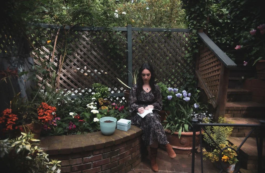 Ashley Tanzosh journaling on a stone bench surrounded by a blooming spring garden during a Liminal Weaver Ostara ritual, embodying seasonal renewal and rebirth
