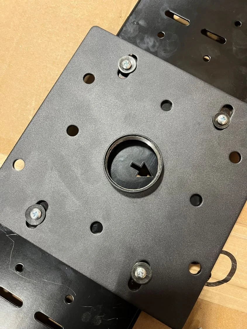 A close-up of a metal mounting plate with a central hole and four screws, lying on a wooden surface.