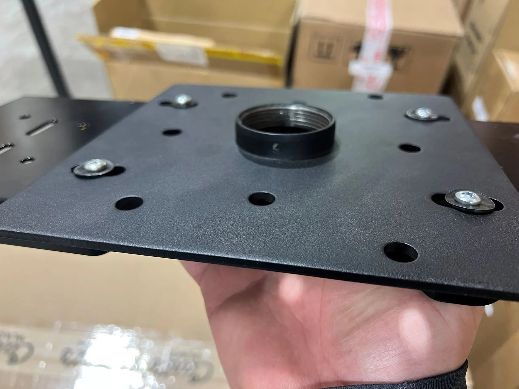 Hand holding a black metal mounting plate with bolts and a central threaded hole, with cardboard boxes in the background.
