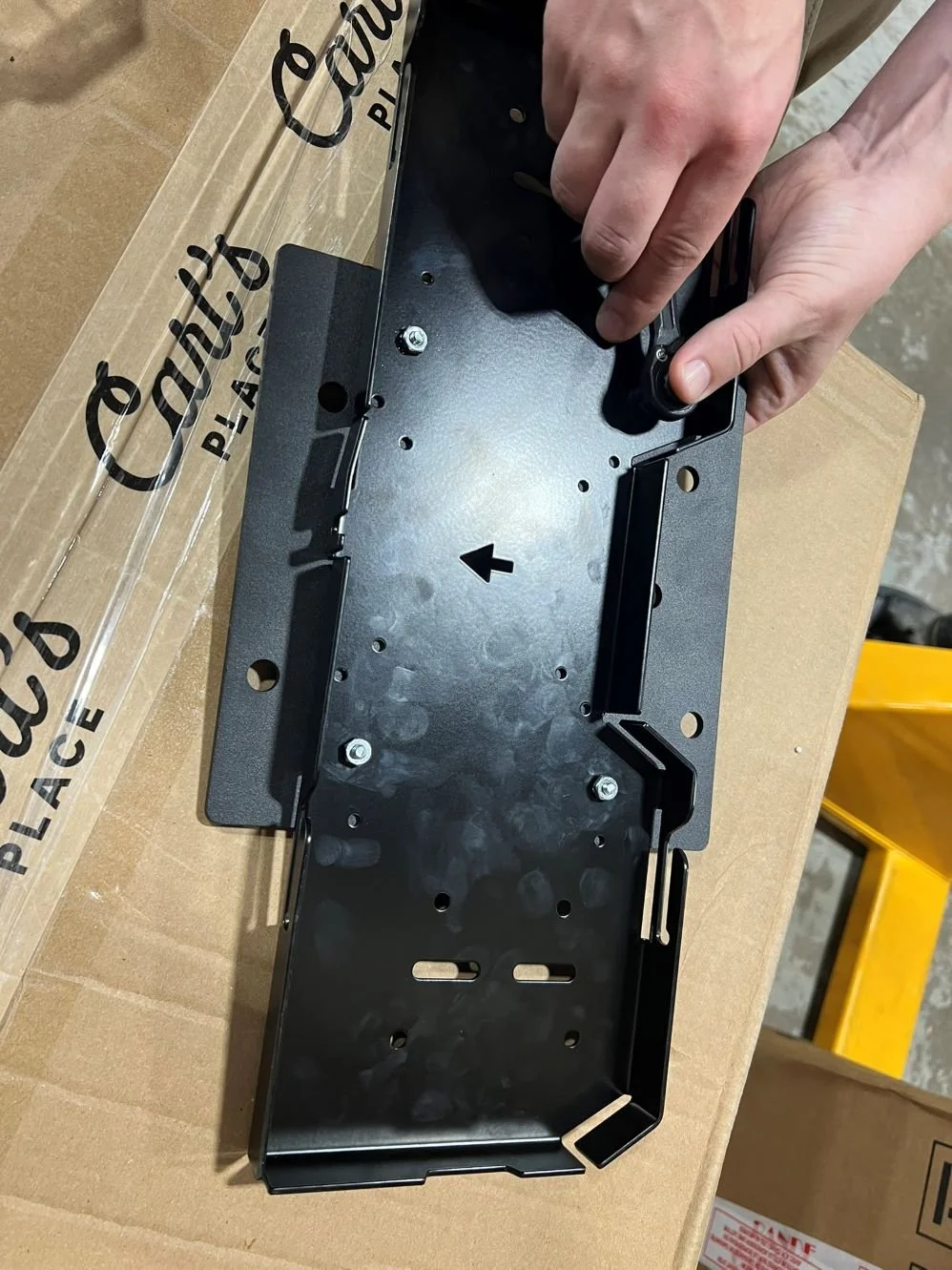 A person assembling or attaching a black metal bracket or tray, possibly part of a larger device or piece of equipment, on top of a cardboard box.