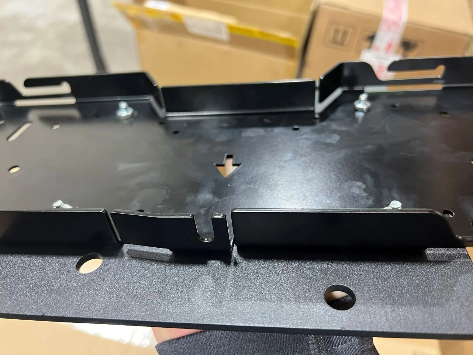 A black metal bracket or mounting plate with several holes and protrusions, held to a surface with screws, with a background of cardboard boxes.