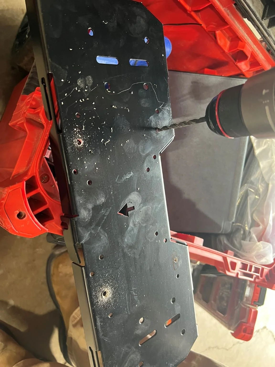 Close-up of a drill bit attached to a black drill, drilling into a metal plate on a workbench.