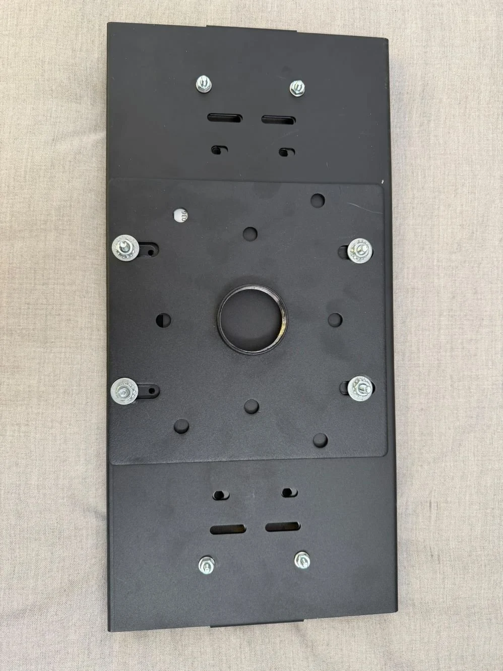 Black metal mounting plate with multiple screw holes and slots, circular opening in the center, and a flat surface, placed on a beige fabric background.