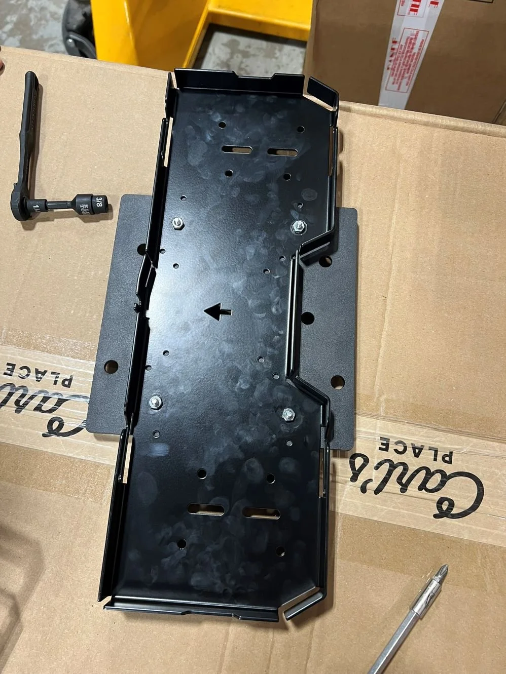 Black metal mounting bracket on a cardboard box, with a small L-shaped tool and a silver pen nearby, in a work area with yellow and cardboard boxes.