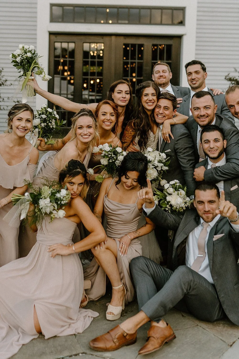A joyful wedding party posing for a group photo. The bridesmaids are wearing light-colored dresses and holding bouquets, while the groomsmen are in gray suits. They are positioned closely together, smiling and celebrating in front of a building with large windows.