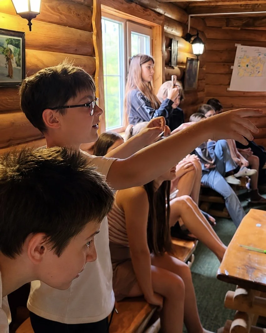 A boy in glasses laughs and points across a wooden room full of children.