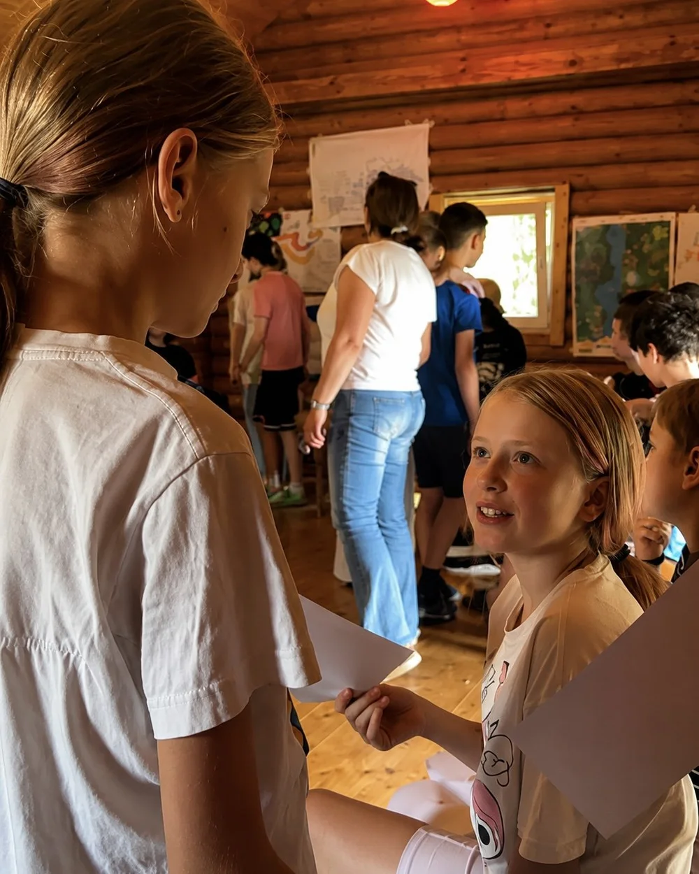Two girls in a log-walled room; one smiles over her shoulder at the camera while other children talk in the background.