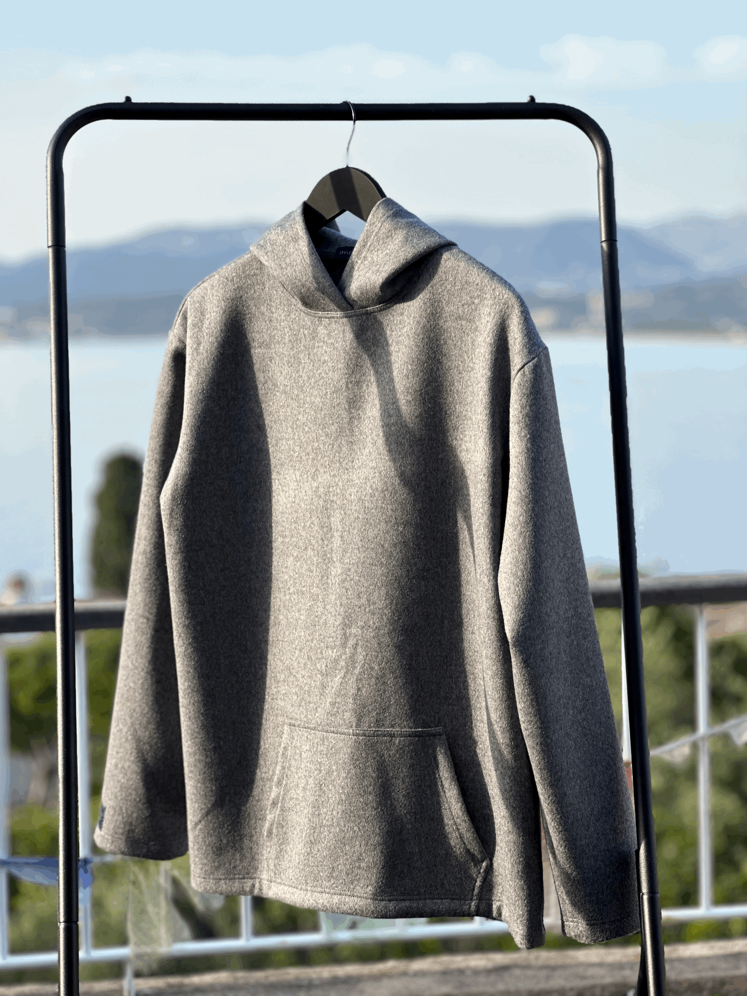 Granite Island Silvery Grey Angora Hoodie hanging on a clothing rail overlooking the sea, displaying its relaxed fit, hood and front pocket.