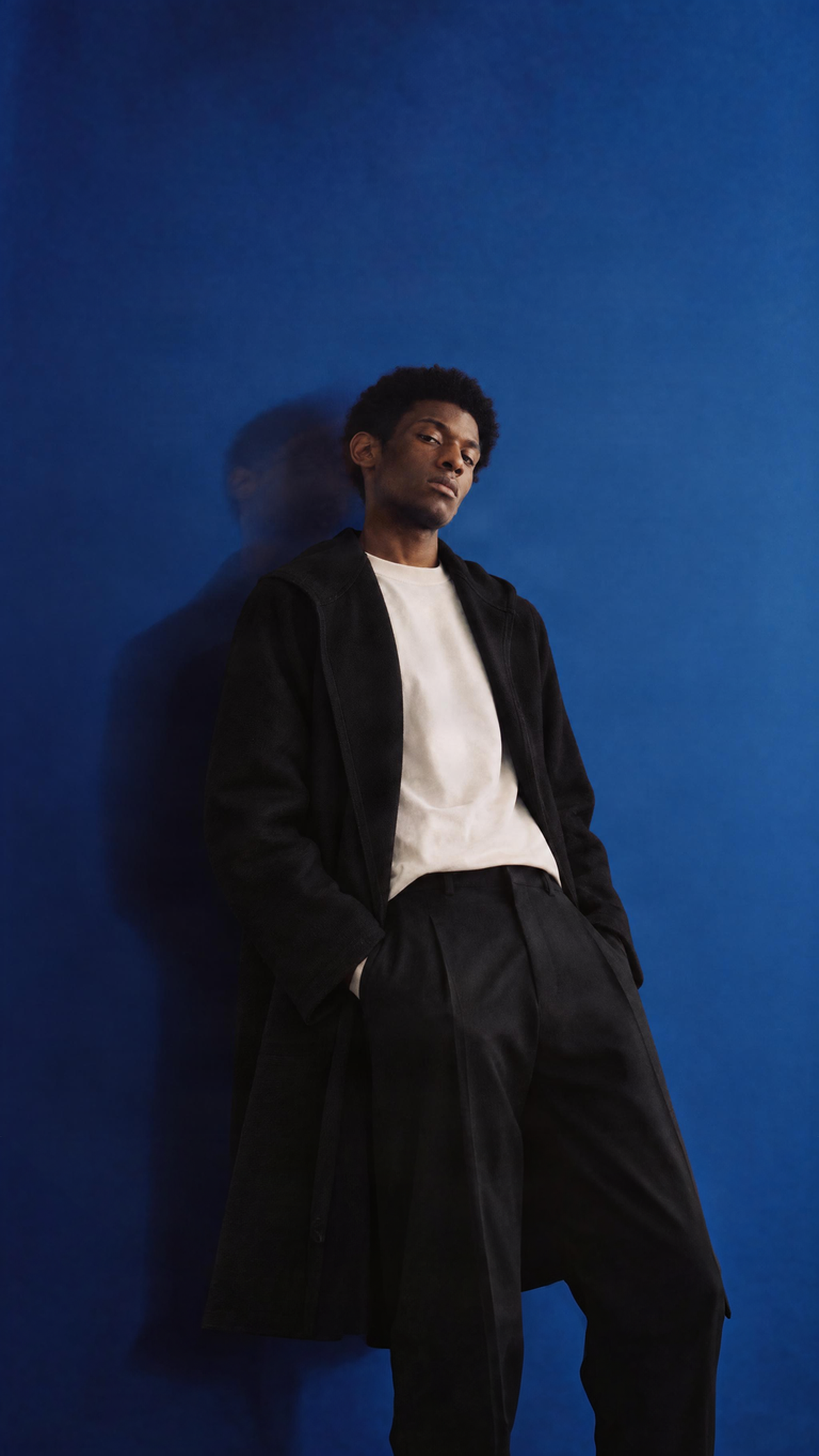 Male model wearing the Granite Island black hooded kimono overcoat, styled with a white T-shirt and tailored black trousers against a blue studio background.